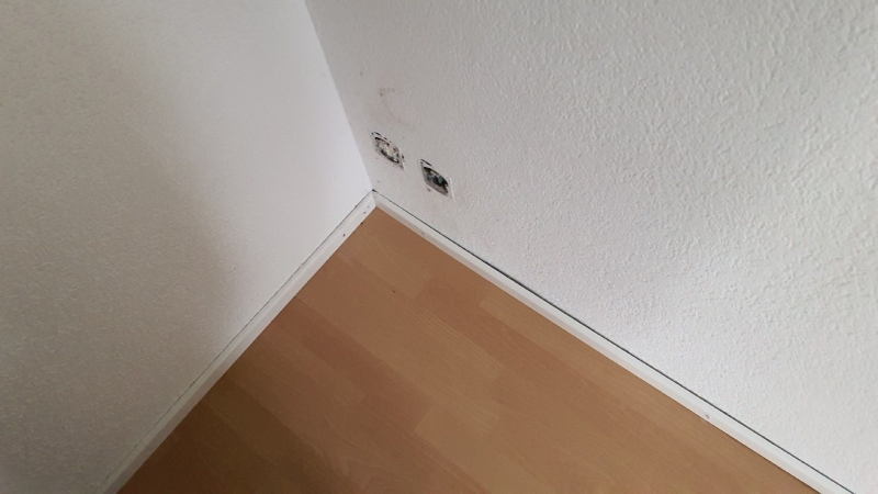 An der schlichten Zimmerecke in Reinach deutet die versteckte Stelle hinter den offenen Steckdosen auf moeglichen Befall durch Bettwanzen hin, weshalb ein Kammerjaeger sinnvoll ist.