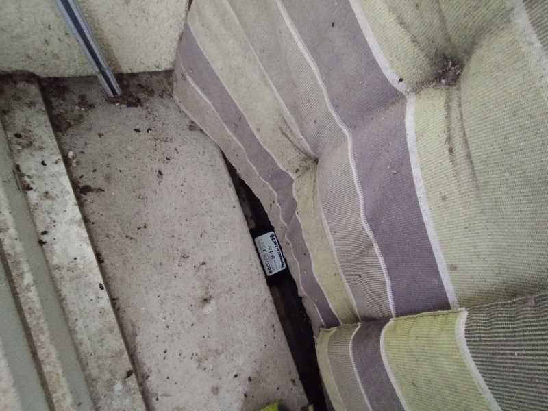 Zwischen verschmutztem Boden und Polsterauflage in Blauen deutet die versteckte Koederbox auf moegliche Maeuseaktivitaet hin, bei der ein Kammerjaeger notwendig sein kann.